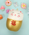 A cute Japanese lucky cat squishy toy with gold and white colors, surrounded by colorful mini doughnut, macaron, and cupcake figurines on a light blue background.