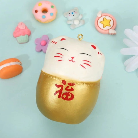 A cute Japanese lucky cat squishy toy with gold and white colors, surrounded by colorful mini doughnut, macaron, and cupcake figurines on a light blue background.