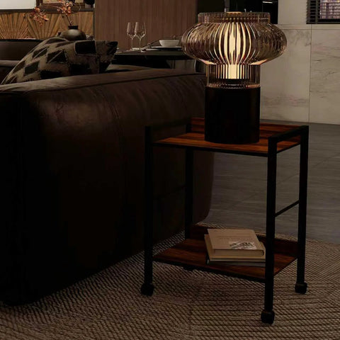 Two Tier Rolling Utility Cart With Brown Wood Shelves And Black Metal Frame On Wheels beside a sofa with decorative lamp on top