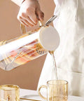 A Game Changer High Borosilicate Glass Pitcher With Secure Tight Lid And Long Mixing Spoon One Point Six Liter Heat Resistant Beverage Carafe pouring tea into a clear glass.