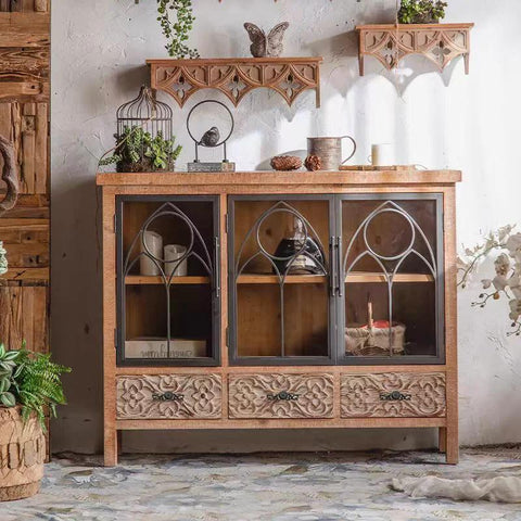 Elegant rustic cabinet with glass doors and intricate carvings, styled with decorative items in a vintage-inspired room.