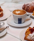 Cup of Lavazza coffee with heart latte art, surrounded by pastries on a table setting.
