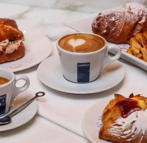 Cup of Lavazza coffee with heart latte art, surrounded by pastries on a table setting.