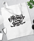 White tote bag with "Always in My Heart" design, sunglasses, greenery, and crafting tools on a gray surface.