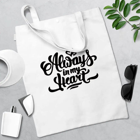 White tote bag with "Always in My Heart" design, sunglasses, greenery, and crafting tools on a gray surface.