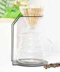 Iron Frame Coffee Dripper Pour Over Stand Nonslip Base with paper filter over a glass carafe and coffee beans.