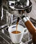 Espresso shot being poured into Lavazza cup from espresso machine with steam wand and wooden handle in a kitchen setting.