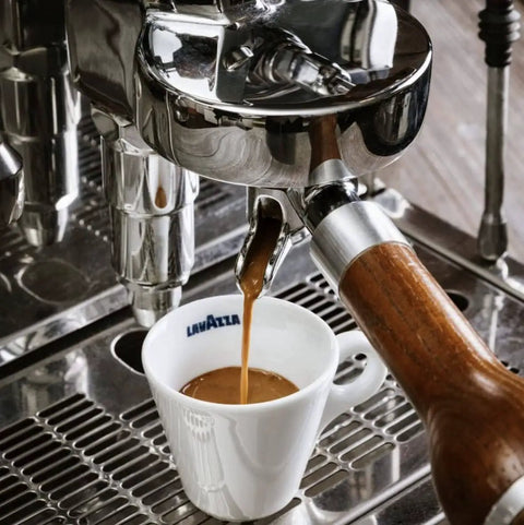 Espresso shot being poured into Lavazza cup from espresso machine with steam wand and wooden handle in a kitchen setting.