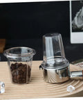Clear espresso dosing cup for coffee machines, anti scatter powder design, next to a portafilter on a wooden base