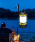 Two Pack LED Camping Lanterns That Are Lit With 1000LM hanging outdoors by a campfire with a person sitting nearby at dusk.