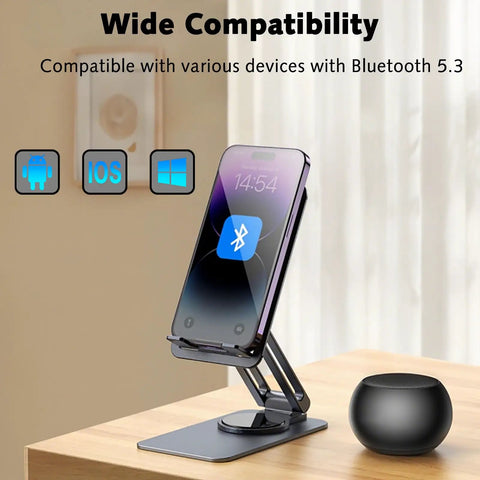 Smartphone on adjustable stand beside pocket-sized Bluetooth speaker with deep bass and crisp highs, 5W stereo sound, on a table.