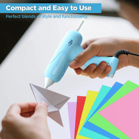 Hand holding Fast Heating Mini Hot Glue Gun Kit With Anti Scald Nozzle, Compact in blue, used on paper crafts with glue sticks.