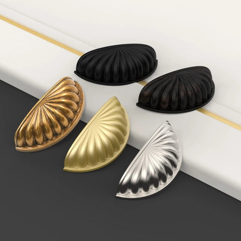 Set of assorted metallic cup pulls on a black and white surface, showing matte black, bronze, gold, and silver finishes for cabinets.