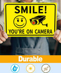 Person holding a Smile You're on Camera sign, 4 Pack Smile Youre On Camera Signs 8x12 Rust Free Aluminum Outdoor Video Surveillance Signs
