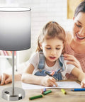 Set Of 2 Bedside Table Lamps For Bedroom And Living Room With 3 Color Temperature Settings USB Charging Port And AC Outlet Pull Chain White Shade Includes 2 Bulbs For Cozy Vibes next to a mother and daughter at a table.