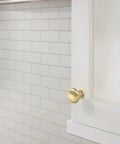 Edona Polished Brass Cabinet Knob 1-1/4 Inch Diameter for cabinets and drawers on white cabinet against tile wall