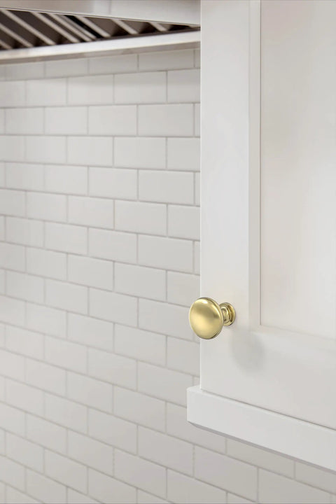 Edona Polished Brass Cabinet Knob 1-1/4 Inch Diameter for cabinets and drawers on white cabinet against tile wall