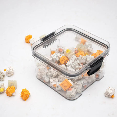 Front-angle view of a clear plastic keyboard switch storage box with a black latch, showing white and orange switches.