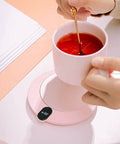 Keep Your Daily Brew On Lock With This Sleek Pink USB Coffee Mug Warmer That Slaps With Three Custom Heat Levels And A Smart Hydration Reminder For Top Shelf Home Office Vibes. A hand stirring tea in a pink mug on a pink USB mug warmer.