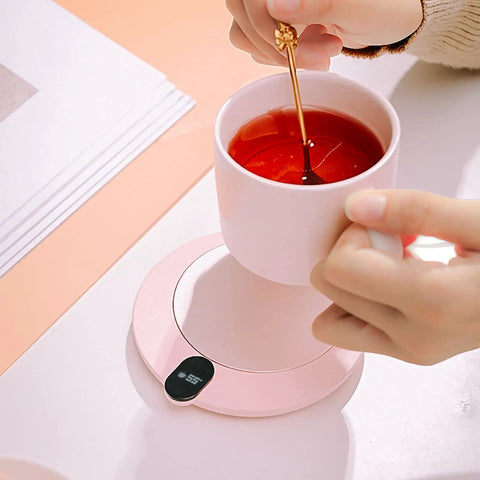 Keep Your Daily Brew On Lock With This Sleek Pink USB Coffee Mug Warmer That Slaps With Three Custom Heat Levels And A Smart Hydration Reminder For Top Shelf Home Office Vibes. A hand stirring tea in a pink mug on a pink USB mug warmer.