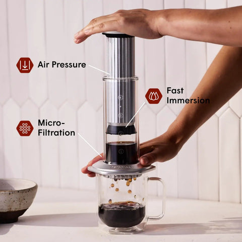 Hand pressing down on All In One Coffee Press With Glass Chamber, Stainless Steel And Aluminum Build, French Press Pour Over And Espresso Style Manual Brewer, 2 Minute Brew For Rich Flavor And Less Bitterness, Silver, atop a clear mug with coffee.