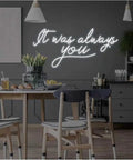 White neon sign "It was always you" over a dining table in a modern kitchen setting.