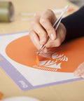 Cricut basic tool set in use; close-up of hands weeding orange vinyl on purple cutting mat