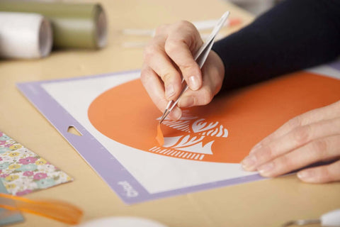 Cricut basic tool set in use; close-up of hands weeding orange vinyl on purple cutting mat