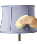 Hand using Easy To Use Sponge Lamp Shade And Fabric Cleaner For Delicate Fabrics And Lampshades, No Fuss Dust, Dirt, And Pet Hair Removal With Dry Magic, No Discoloration Risk, Hand Wash, Air Dry, And Reusable Clean In Between on a purple lampshade.