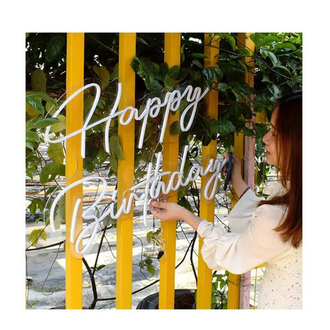 Woman holding Neon LED Birthday Sign USB Powered Acrylic Wall Light For Party Vibes Teen Kids Bedroom Living Room Bar Mitzvah Gift Prom 16.3x10.2 Inches Dreamy Glow For Any Party Or Holiday Easy USB Setup against yellow fence with greenery.