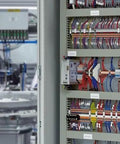 Image showing internal view of an electrical control panel with wires and components. Background includes industrial machinery.