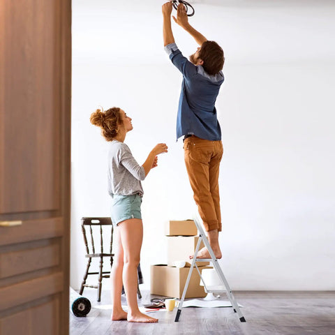 Man standing on Two Step Folding Ladder With Handgrip And Wide Anti Slip Pedals Heavy Duty Safe Lightweight Step Stool for hanging a light fixture.