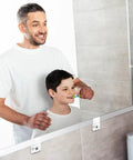 A father helps his son brush teeth in front of a mirror secured with Pretty Slick Mirror Holder Clips With Screws For Effortless Frame And Cabinet Door Mounting Includes Thirty Clear Plastic Hanging Hardware Sets For Quick Wall Glass Install Jobs.
