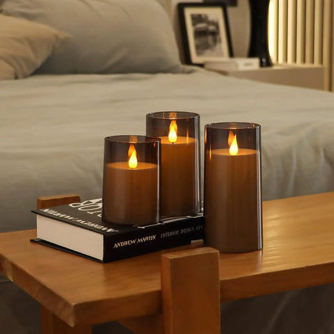 Grey Flameless Pillar Candles With Remote Control And Timer Battery Operated LED Candles Featuring Realistic 3D Flame Perspex Construction Pack Of 3 In D 3 Inch By H 4 Inch 5 Inch And 6 Inch Heights displayed on a wooden table, creating a cozy ambiance in a bedroom setting.