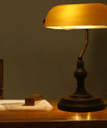 Jade Green Glass Vintage Banker Desk Lamp With Antique Brass Finish on wooden table in dim room, showcasing retro mid century vibe.