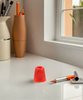 Red cap object on desk beside pens and pencils in a holder by a window.