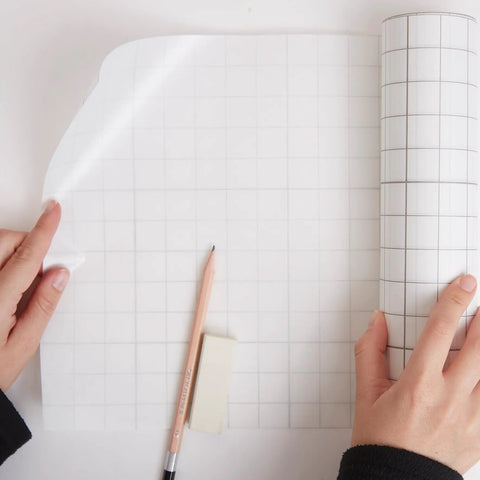 Frisket Masking Film Matte 10 Inch By 4 Yard Roll in use, artist applying film with pencil and eraser on grid surface
