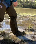 Person wearing Tough As Nails Beefed Up Steel Toe Rubber Boots With Snake Proof Hip Waders in muddy water.