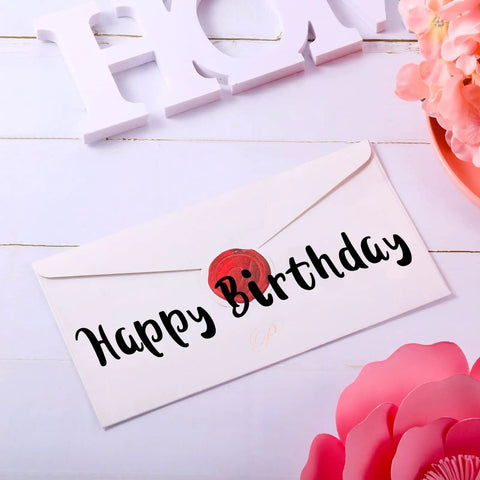 White envelope with 'Happy Birthday' text in black vinyl letters, surrounded by pink flowers, on a white wooden background.