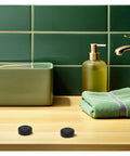 Bathroom sink with two black round objects resembling caps near faucet and towel, against green tiled wall.