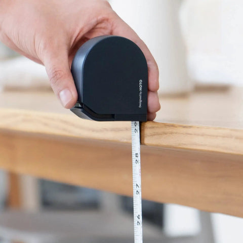 Hand holding navy blue Self Locking Tape Measure Pocket Size Retractable Dual Unit Feet Meters Gadget measuring table edge.