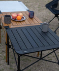 Ultra Lightweight Stable Folding Aluminum Table With Roll Up Top And Carry Bag For Camping Picnics Beach Chill Backyard Barbecues Tailgating And RV Trips Anywhere shown with black and wooden finish, holding mugs and fruit.
