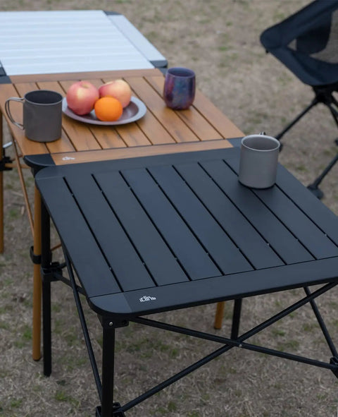 Ultra Lightweight Stable Folding Aluminum Table With Roll Up Top And Carry Bag For Camping Picnics Beach Chill Backyard Barbecues Tailgating And RV Trips Anywhere shown with black and wooden finish, holding mugs and fruit.