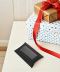 50 Pack Black Kraft Paper Pillow Gift Boxes With Window For Birthday Parties And Events displayed on a table with wrapped gifts.