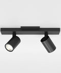 Ascoli Twin Indoor Spotlight Bar In Matte Black Finish With Dual Adjustable Heads Tilt And Rotate For Kitchen Islands Great For Tight Spaces Dry Rated GU10 Lamp Space Saving Ceiling Light Designed In Britain 3 Year Warranty with two cylindrical lights on a rectangular mount in a white room.