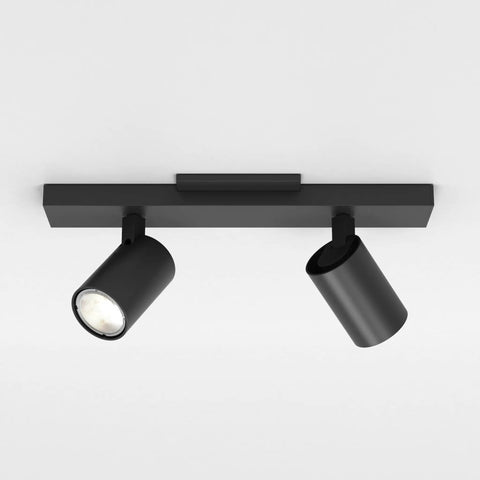 Ascoli Twin Indoor Spotlight Bar In Matte Black Finish With Dual Adjustable Heads Tilt And Rotate For Kitchen Islands Great For Tight Spaces Dry Rated GU10 Lamp Space Saving Ceiling Light Designed In Britain 3 Year Warranty with two cylindrical lights on a rectangular mount in a white room.