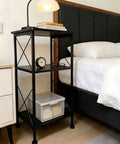 3-Tier Black Storage Shelf With Industrial X-Frame Design next to bed, featuring books, clock, and storage box.