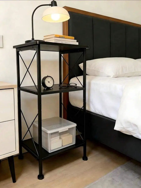 3-Tier Black Storage Shelf With Industrial X-Frame Design next to bed, featuring books, clock, and storage box.
