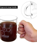 Hand holding 2 Pack 16 Oz Caffeine Beaker Mug Set made of borosilicate glass with realistic caffeine molecule design and ergonomic handle.