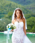 Bride wearing a white lace dress and a rosary necklace, holding a bouquet, standing outdoors with greenery in the background.